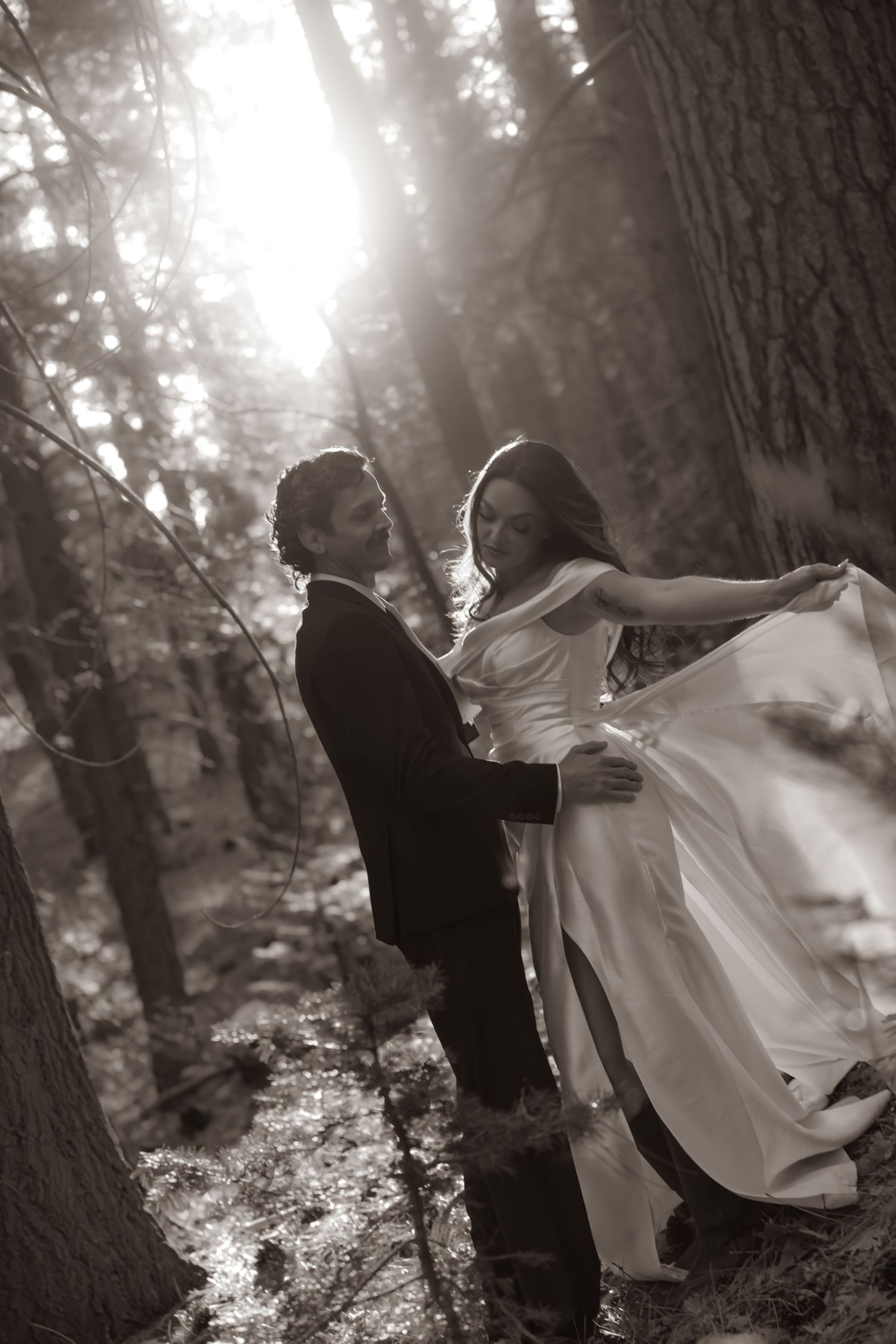 Bride and groom embracing in sunlit forest, sepia-toned wedding photo with flowing white dress.
