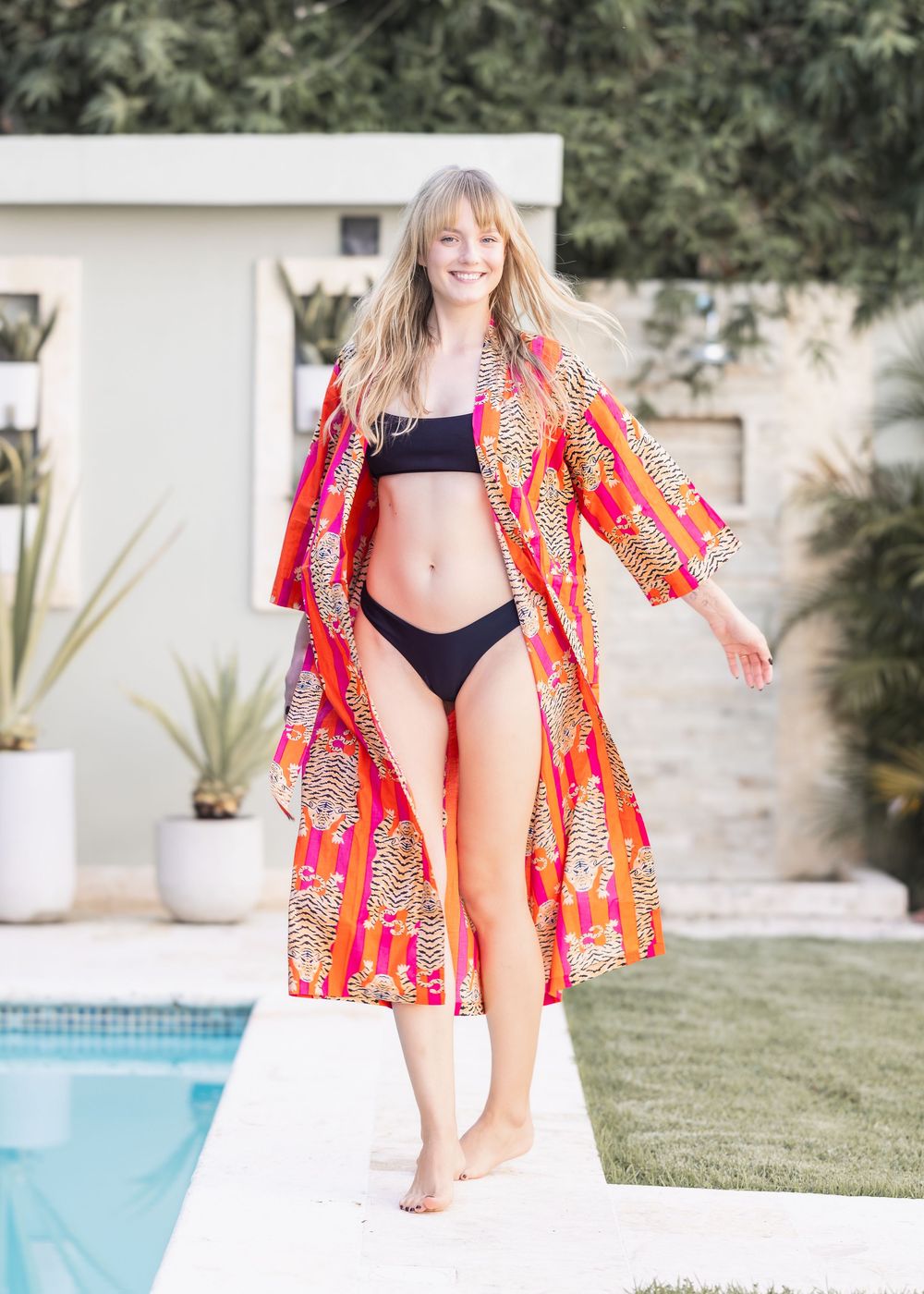 Woman in black bikini and vibrant orange tiger-print kimono cover-up posing by a pool on a sunny day.