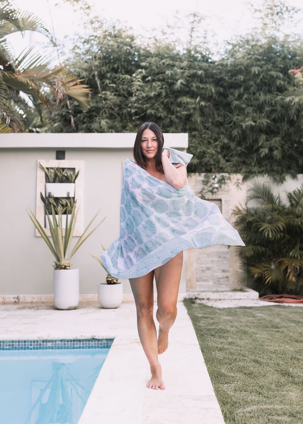 Brunette woman wrapped in a light blue printed beach towel walking by a pool.