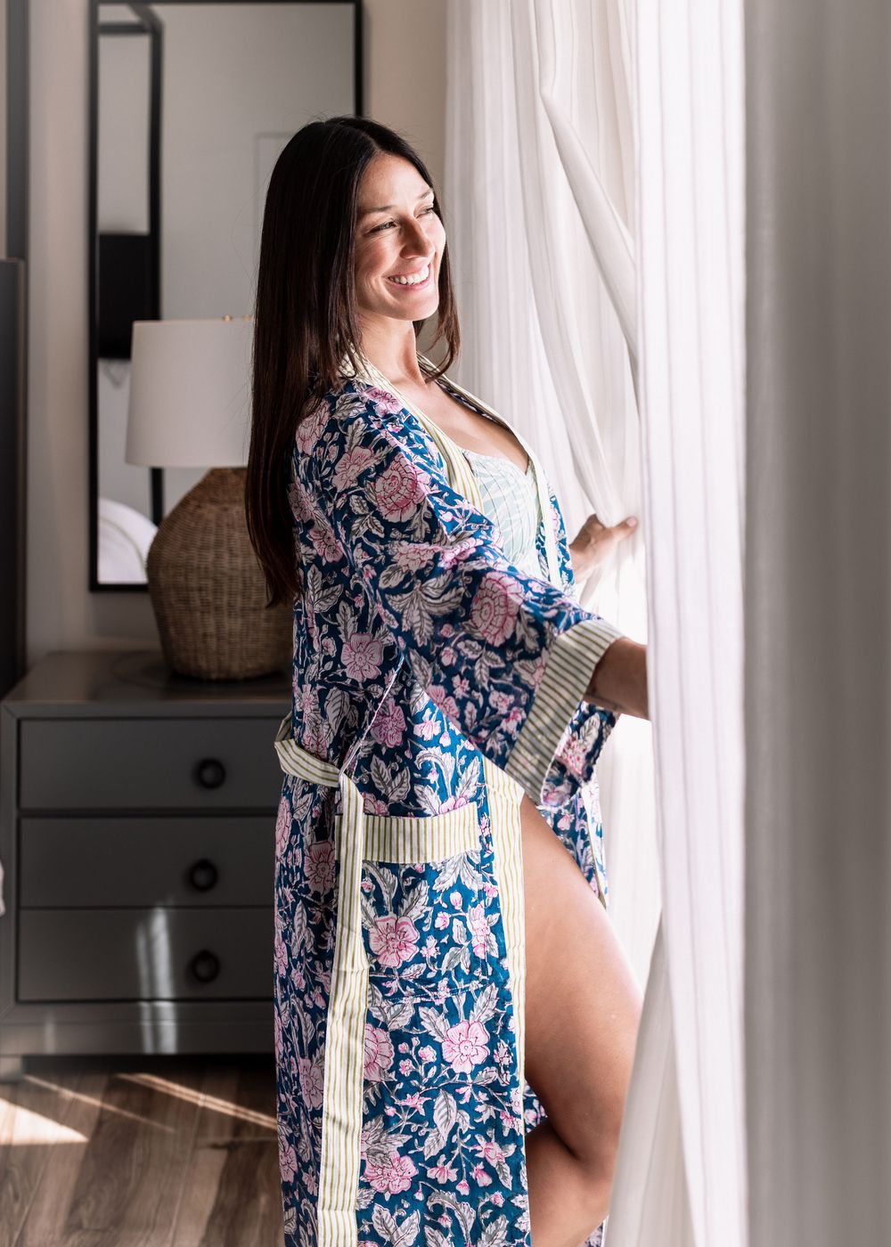 Woman in floral blue and pink kimono robe standing by sheer curtains in a sunlit bedroom.