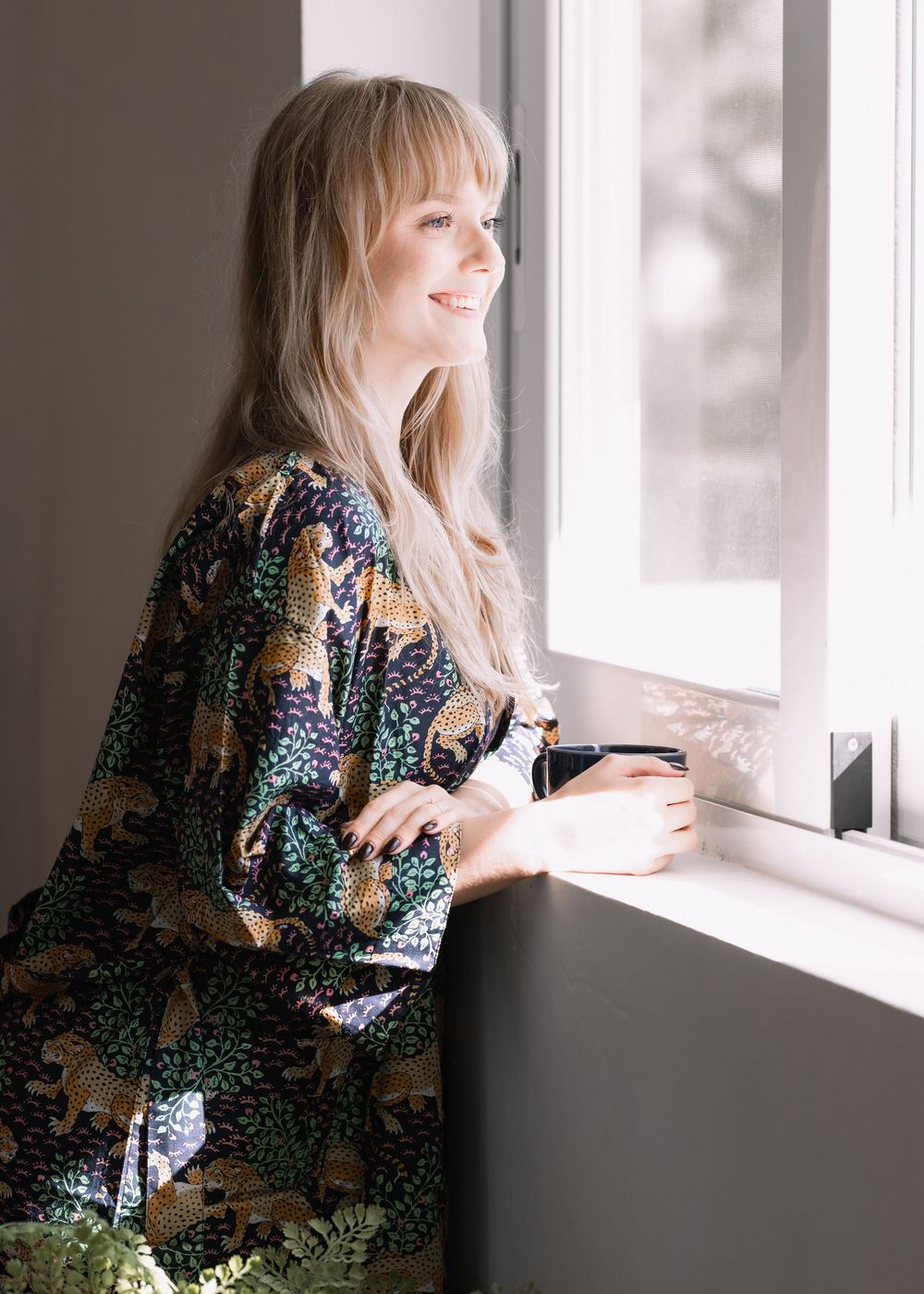 Smiling blonde woman in a dark floral kimono robe gazing out a bright window.