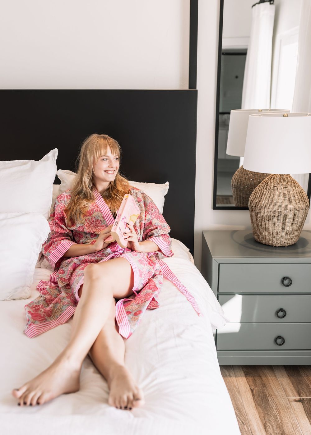 Smiling blonde woman sitting on a white bed wearing a pink floral kimono robe.