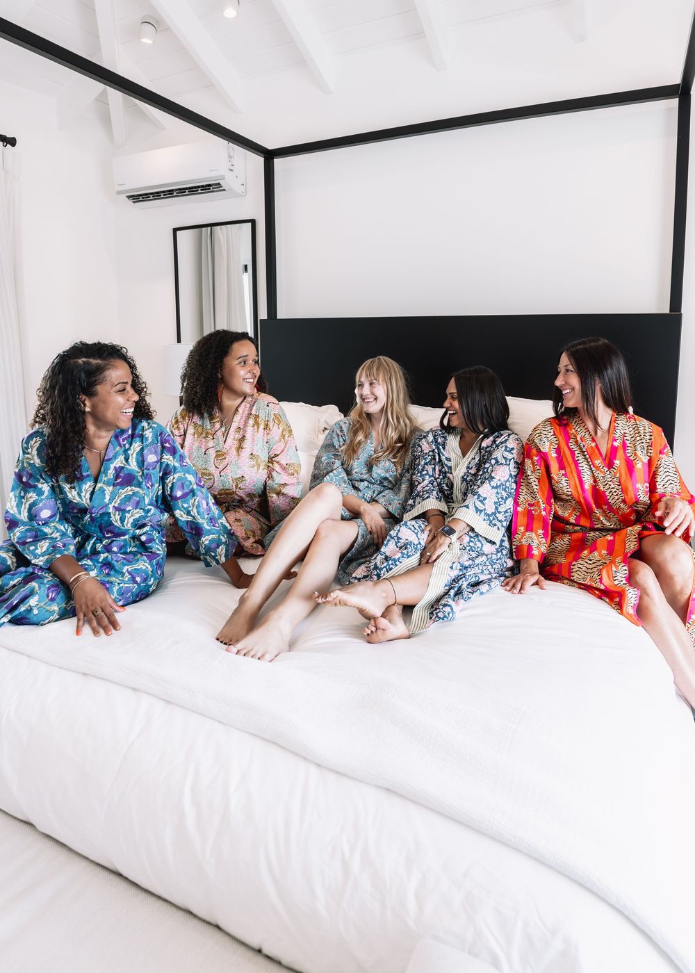 Five women in colorful floral robes sitting together on a white bed, laughing and enjoying a fun girls gathering.