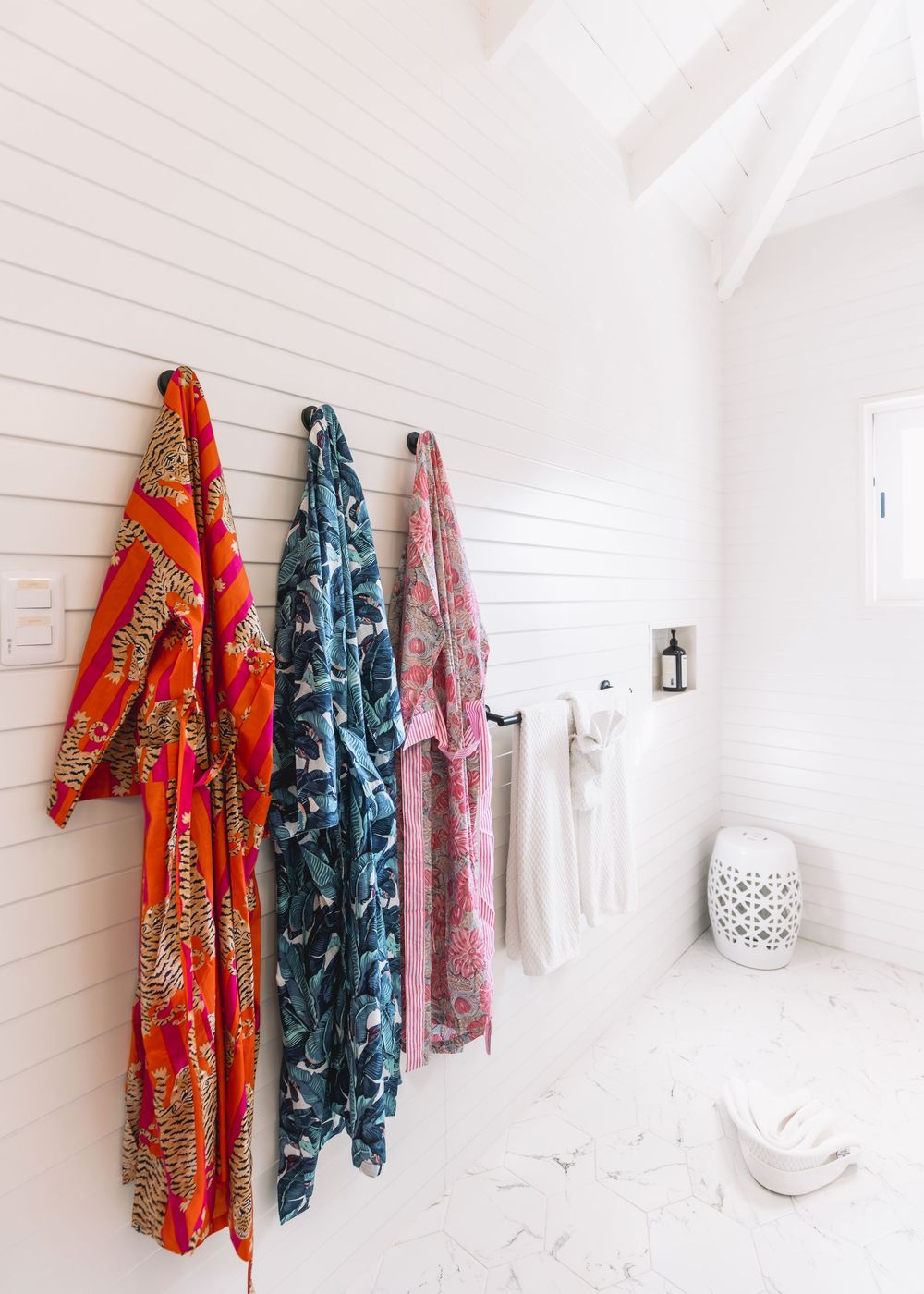 Three colorful printed kimono robes hanging on hooks in a bright white bathroom.