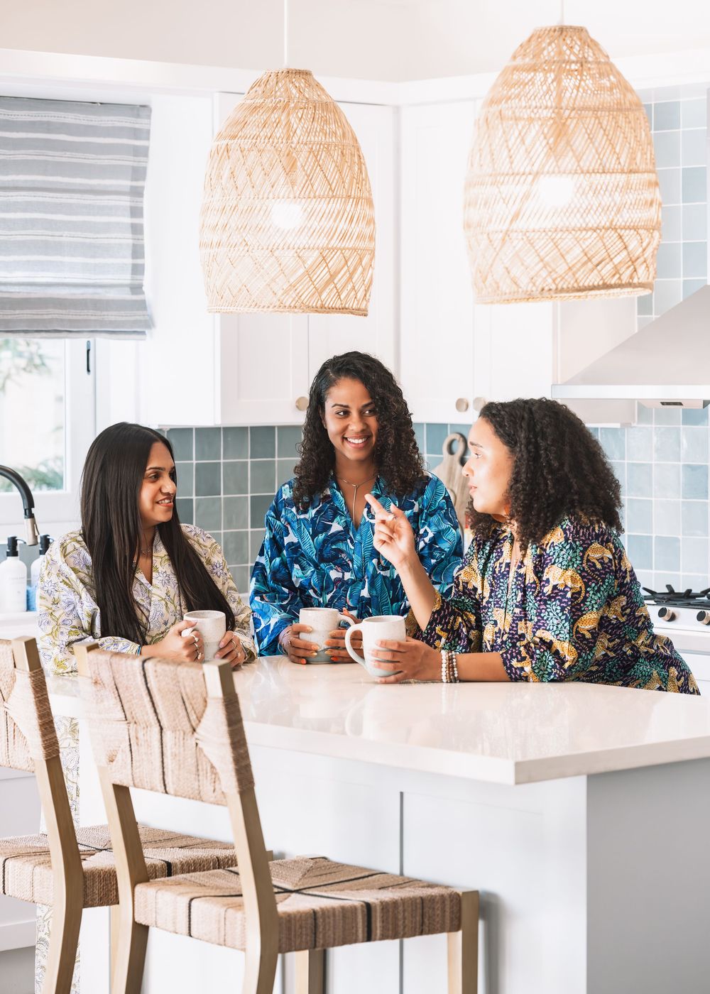 Three women in colorful robes chat over coffee at a bright white kitchen island with wicker pendant lights.