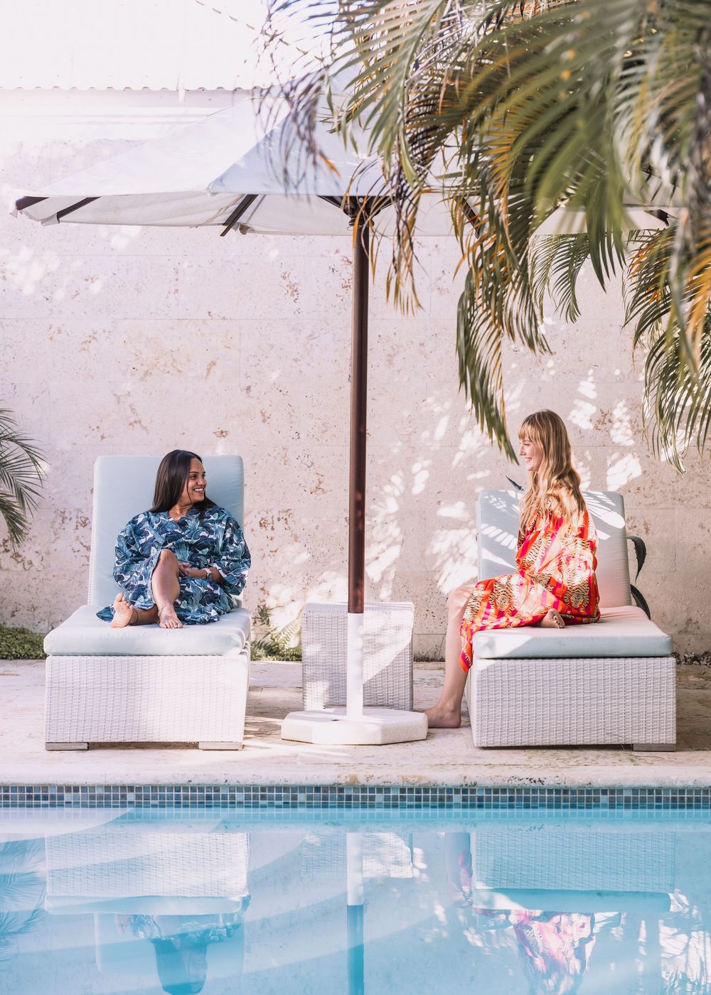 Two women in floral robes relaxing on white lounge chairs beside a pool under a palm umbrella.