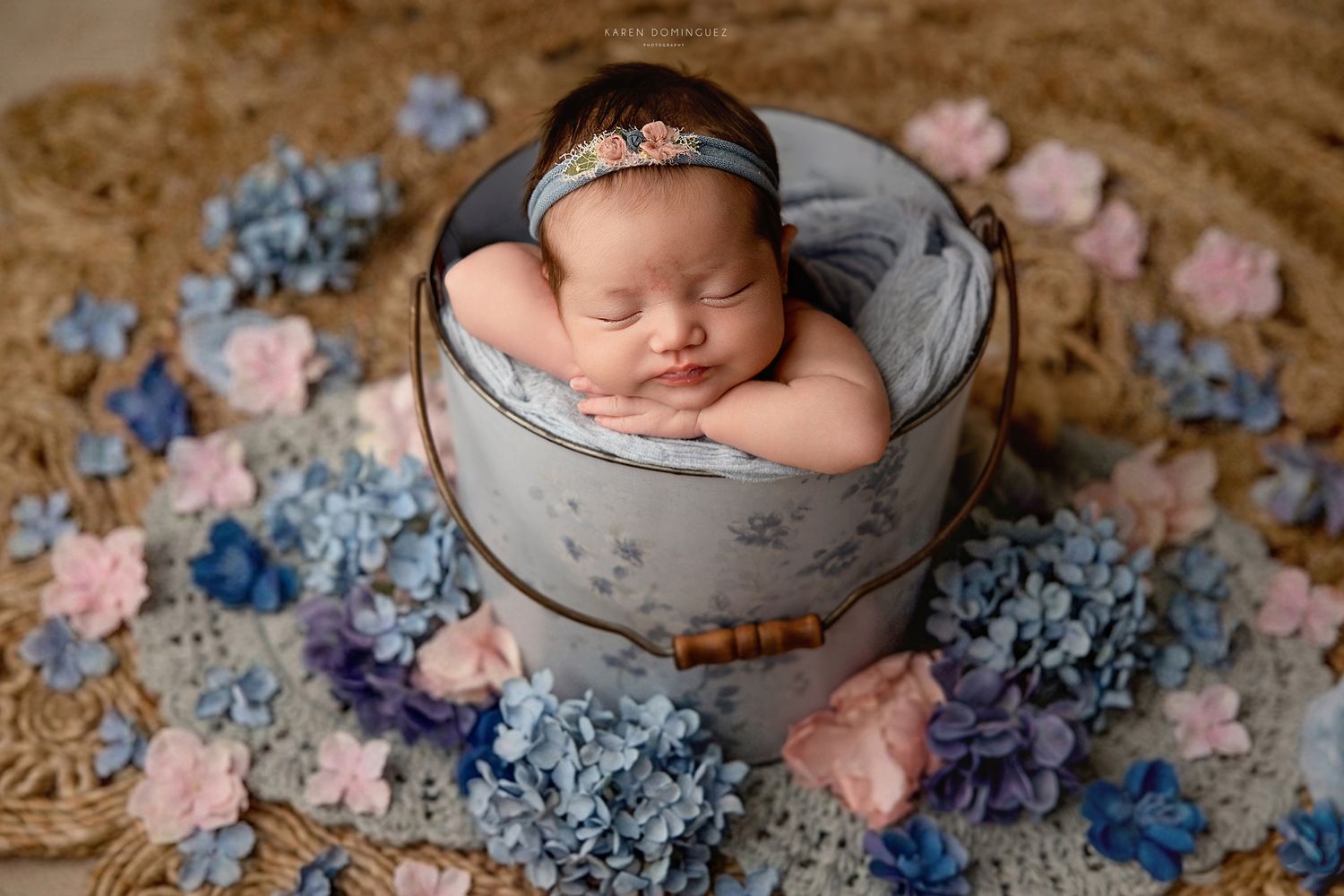 Newborn baby sleeping peacefully in a vintage bucket surrounded by blue and pink hydrangeas on a woven mat.