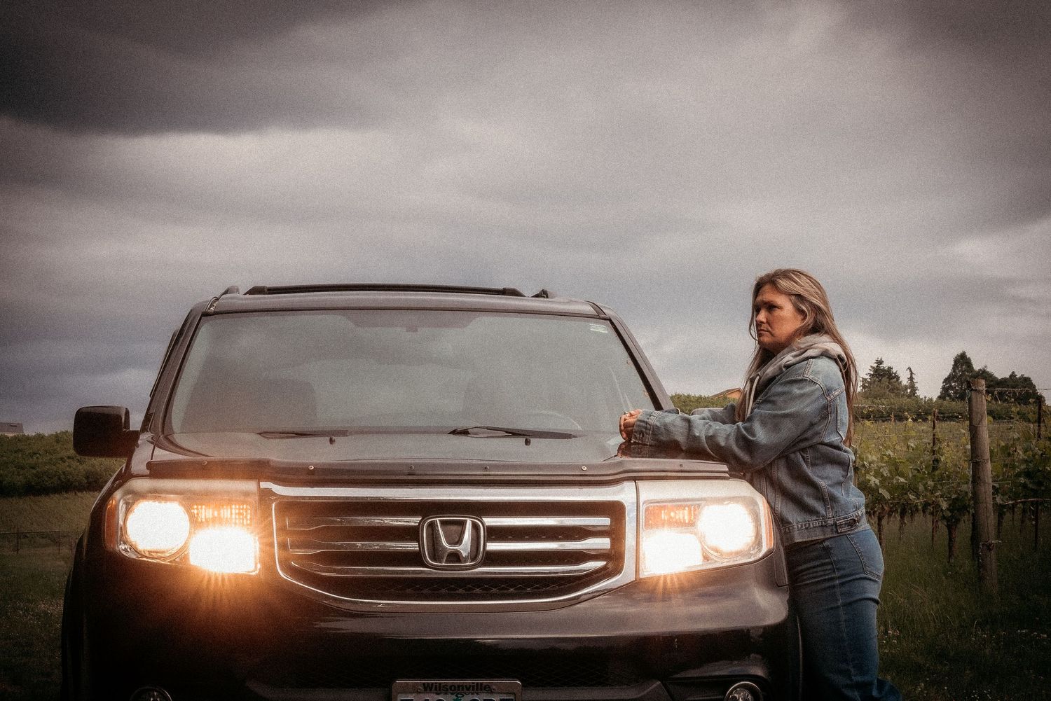Woman leaning on a dark Honda SUV with headlights on, standing beside a vineyard under a cloudy sky.