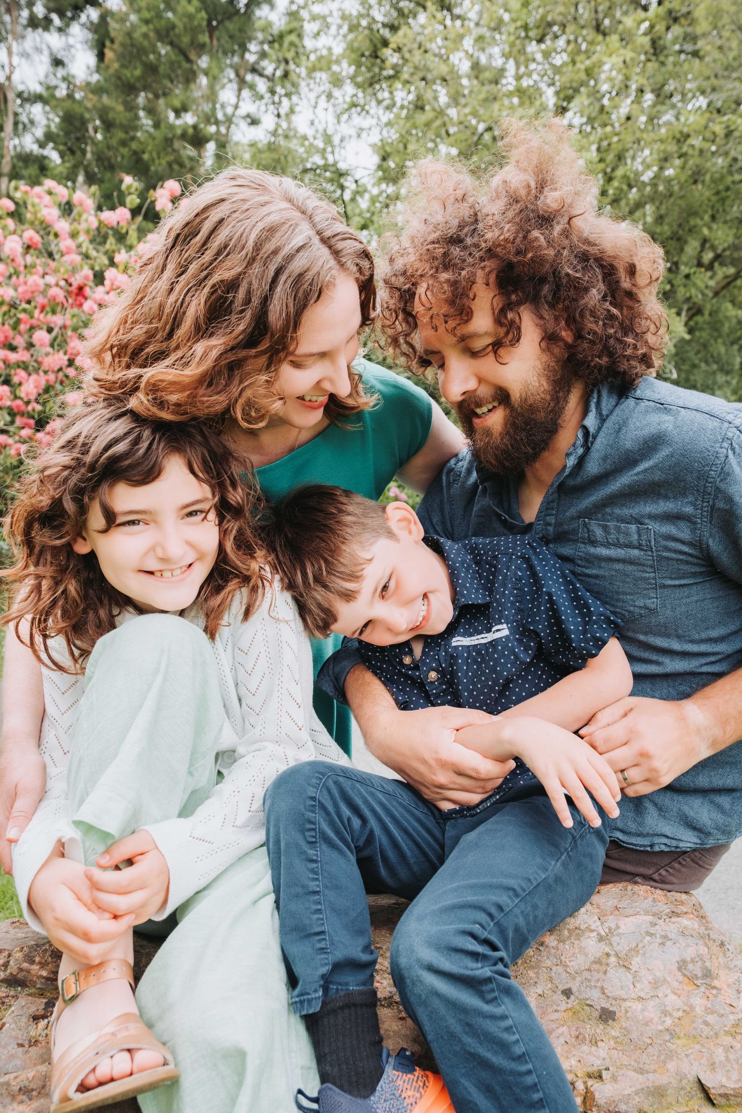 Happy family of four laughing together outdoors, with parents embracing two curly-haired children near blooming pink flowers.