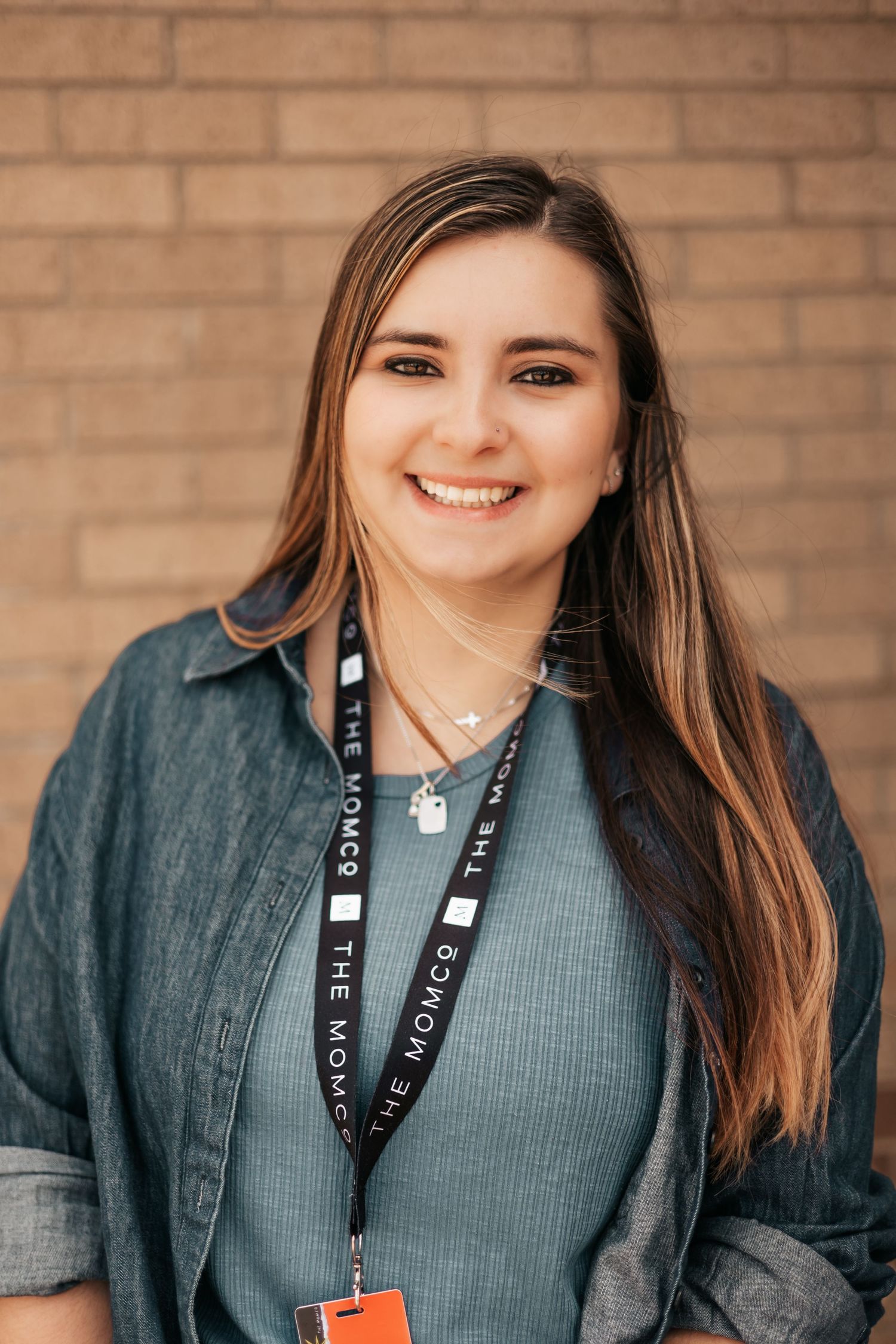 Smiling young woman with long brown hair wearing a denim shirt and lanyard, standing in front of a brick wall.