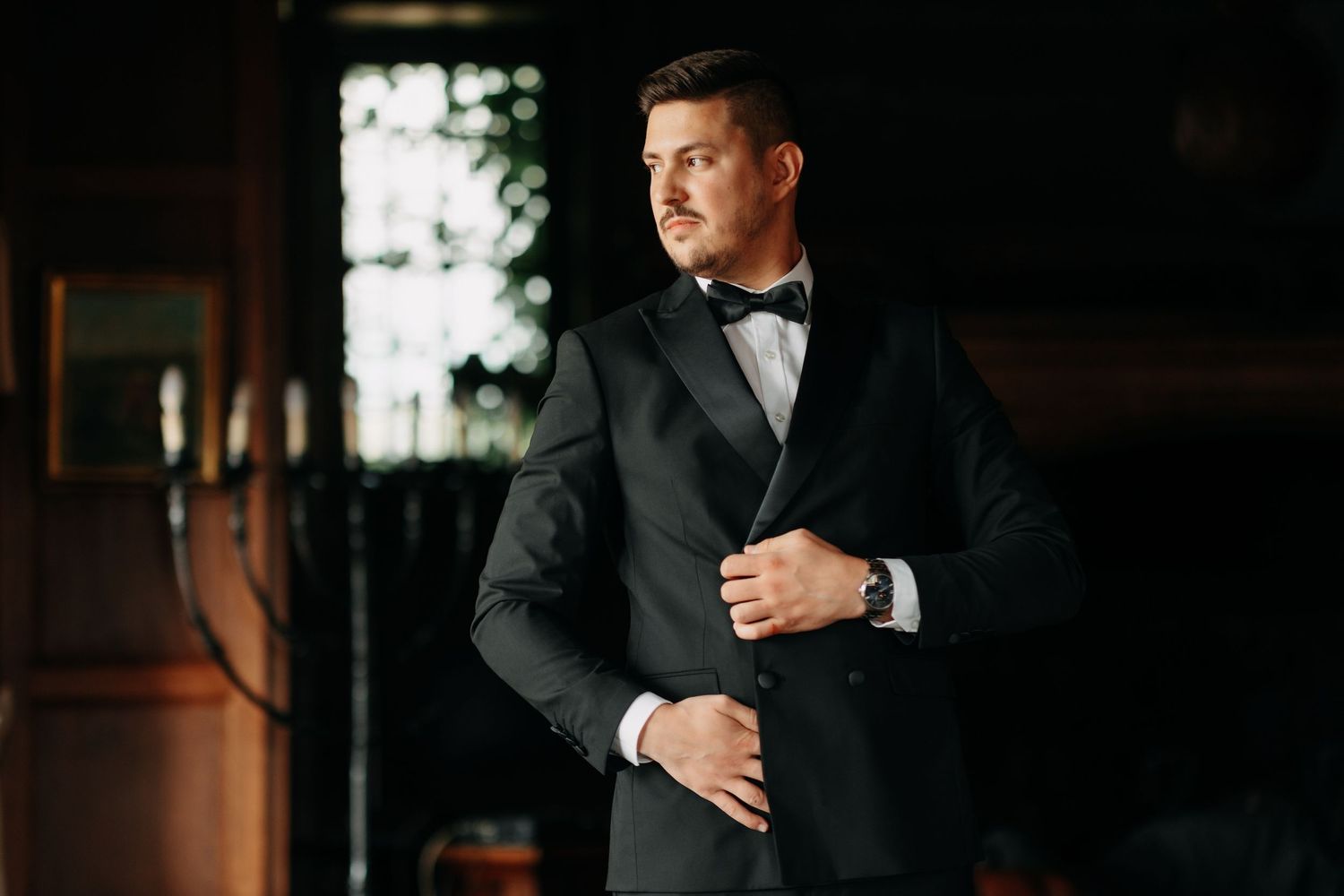 A dapper groom in a black tuxedo and bow tie adjusts his jacket in a dimly lit elegant room.