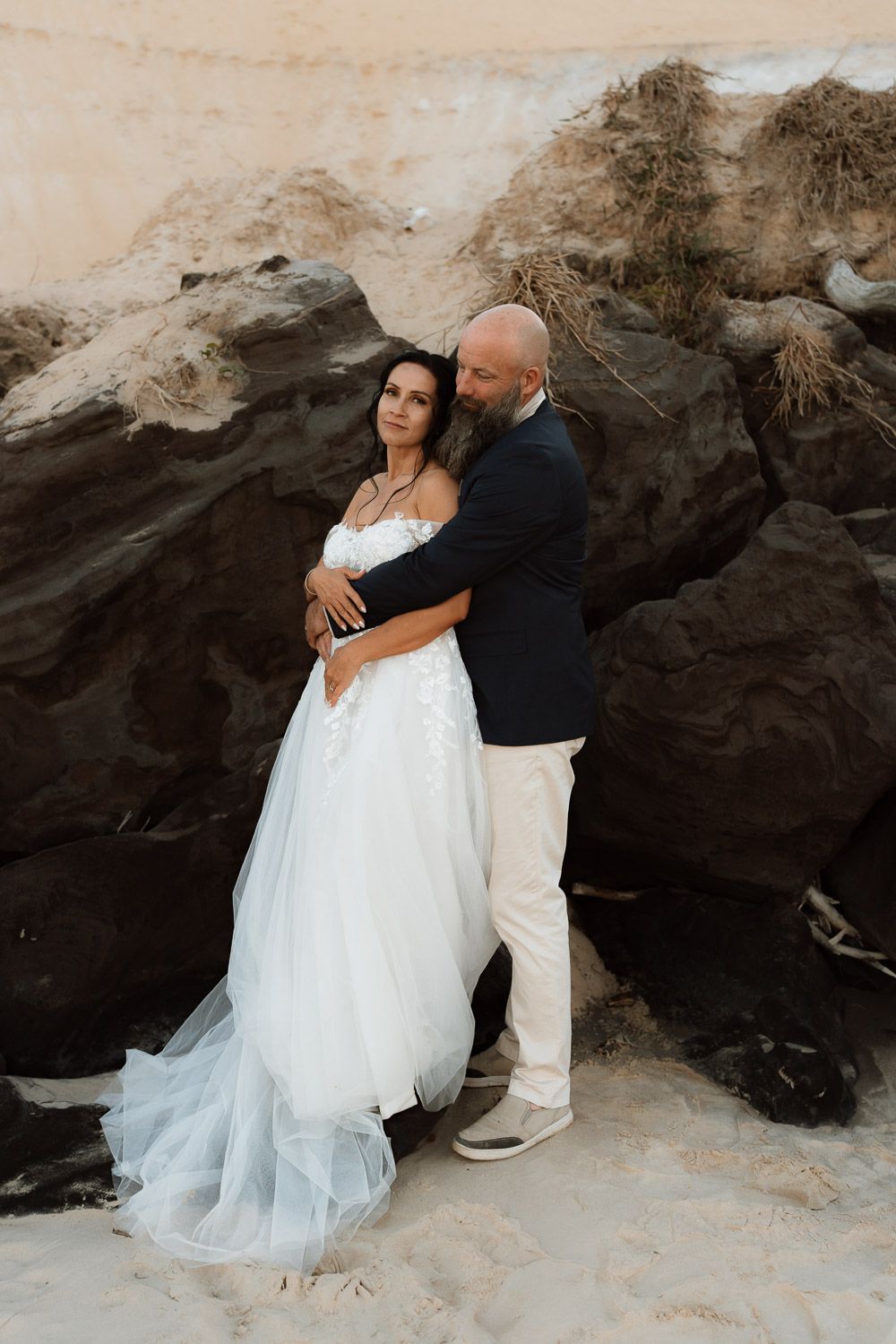 Bride in white gown and groom in navy suit embrace against dark rocks on sandy beach in romantic wedding photo.