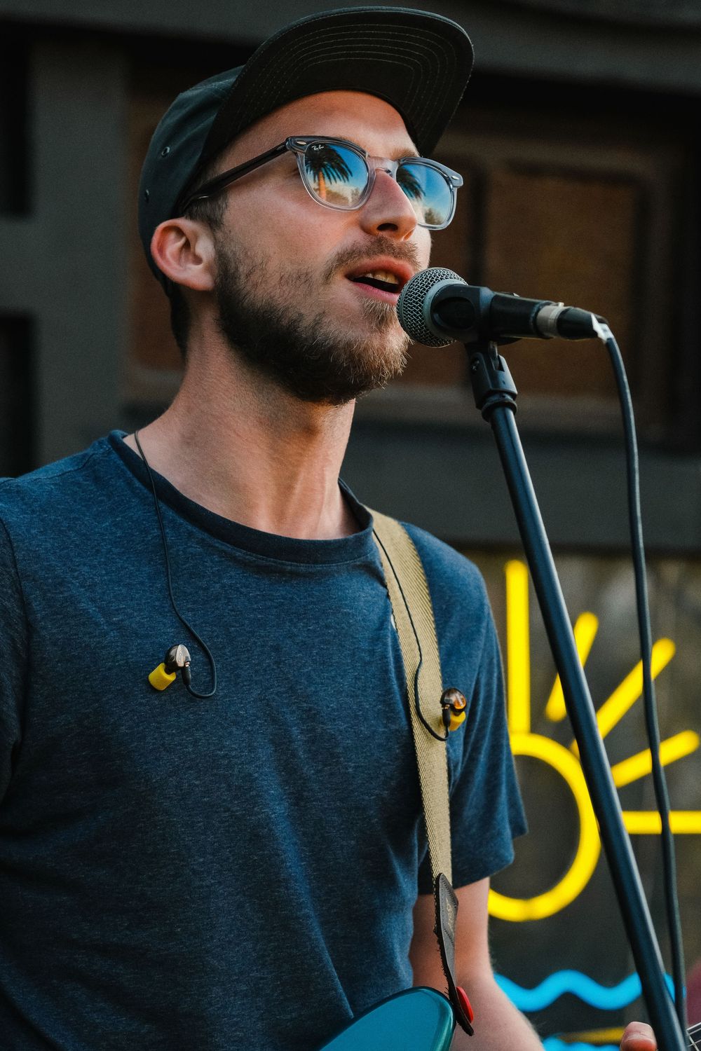Young male musician with beard and cap singing into a microphone while playing guitar outdoors.