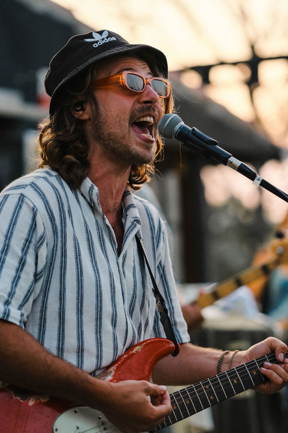 Man with long hair, bucket hat, and sunglasses singing into microphone while playing red electric guitar outdoors.