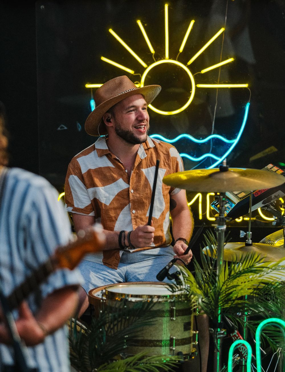 Smiling drummer in tan hat and brown shirt playing cymbals in front of neon sun sign indoors.