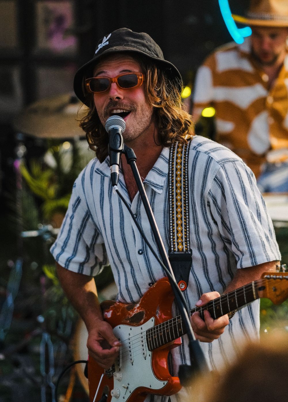 Long-haired guitarist in striped shirt and bucket hat singing into microphone while playing Stratocaster guitar.