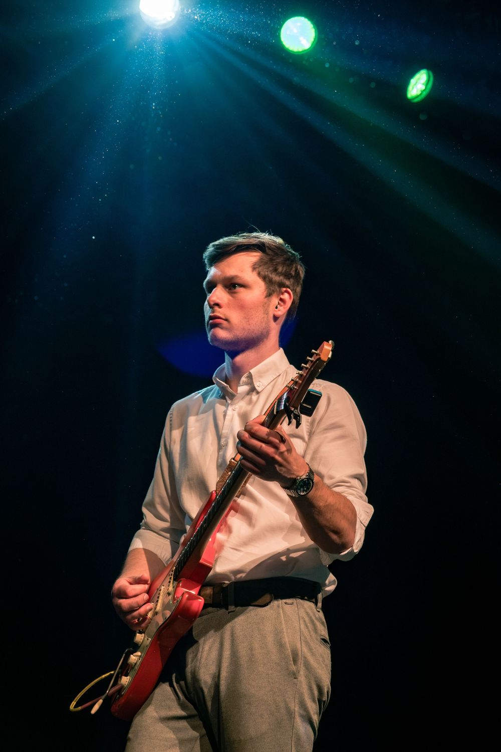 Young male guitarist in white shirt playing red electric guitar under blue and green stage lights.