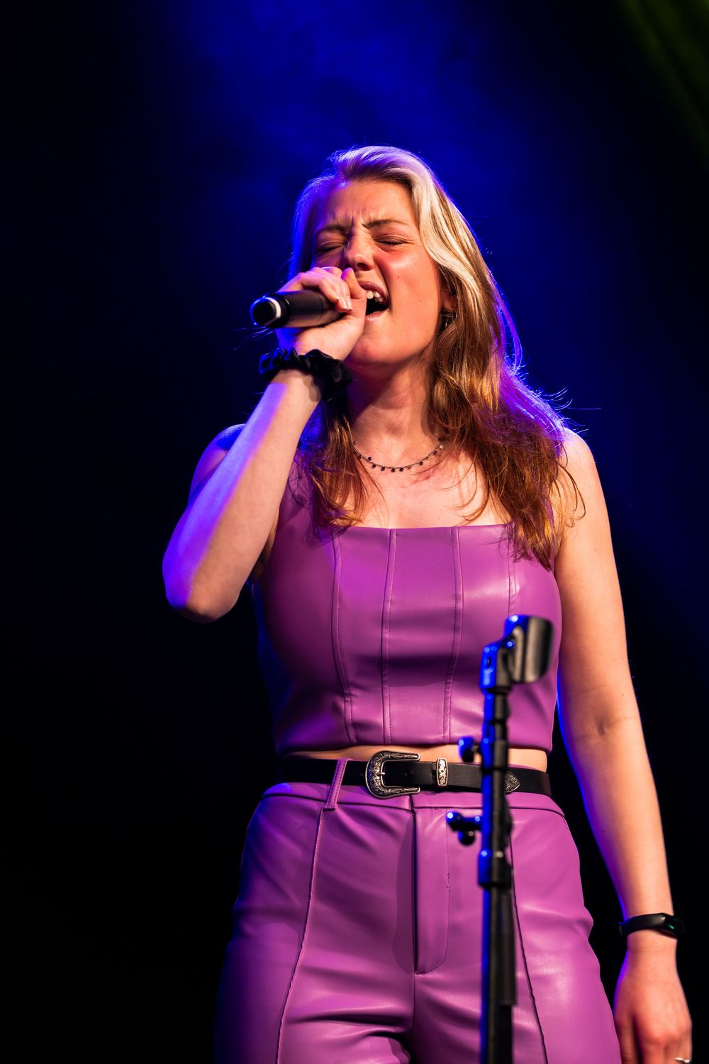Young woman in purple leather outfit singing passionately into handheld microphone on stage.