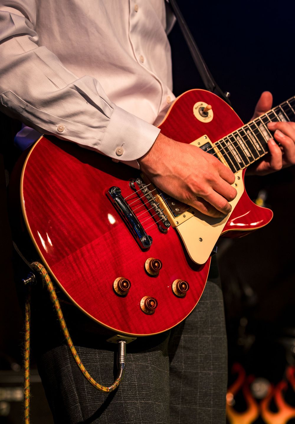 Close-up of hands playing a red Gibson Les Paul electric guitar on stage with flames backdrop.