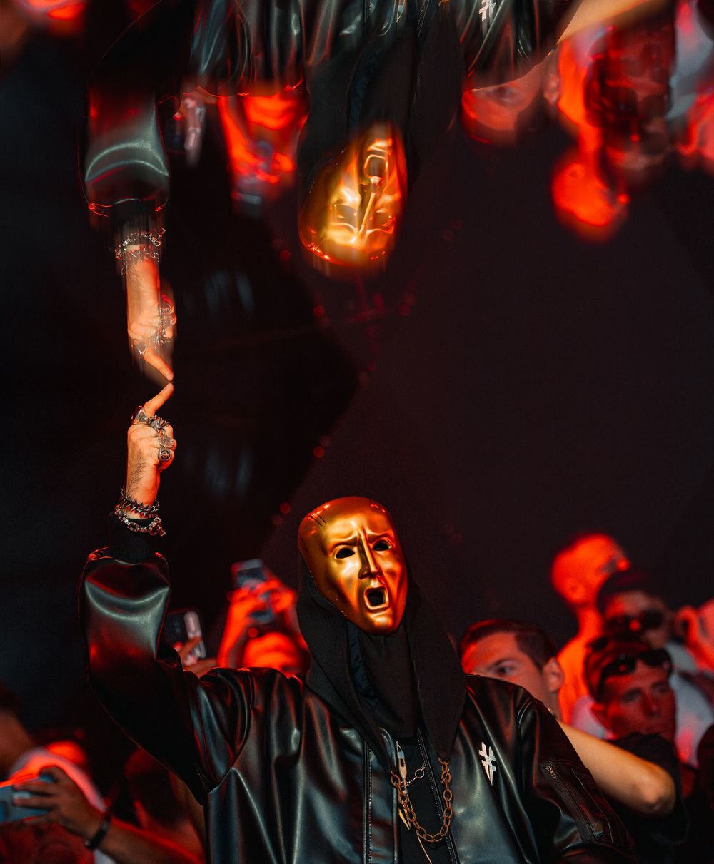 Performer wearing a gold theatrical mask under dramatic red lighting at a dark nighttime music festival.