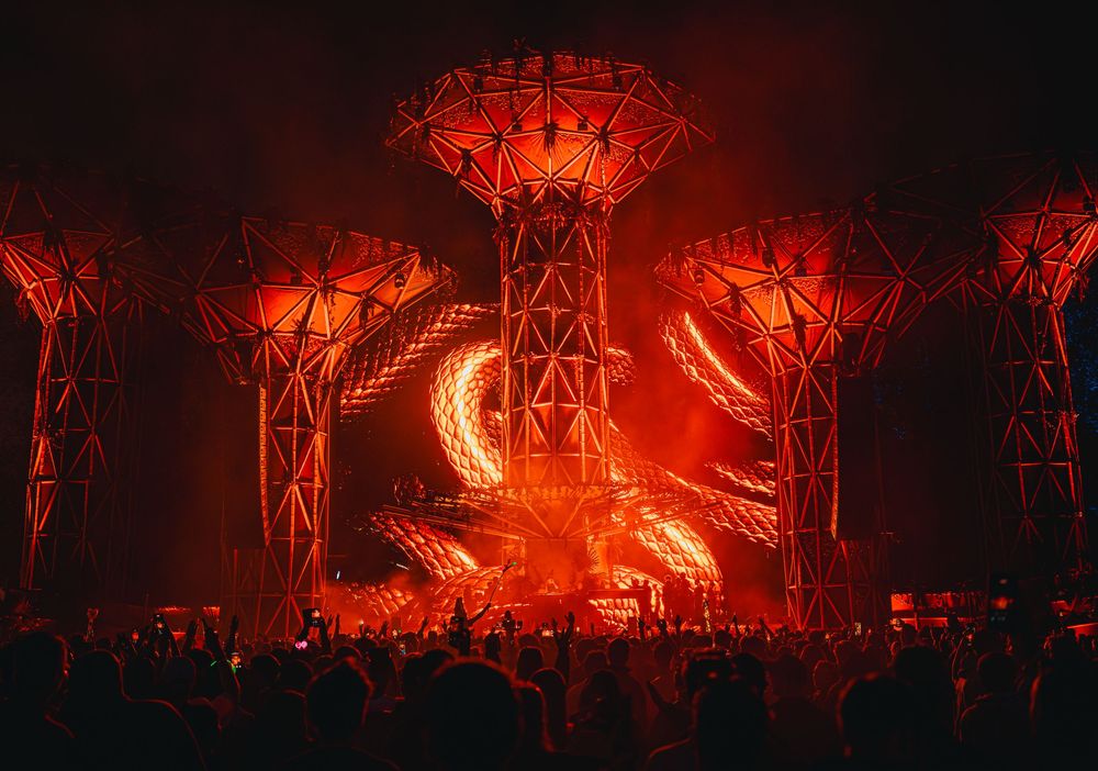 Red-lit outdoor music festival stage with dramatic tree structures and laser light show at night.