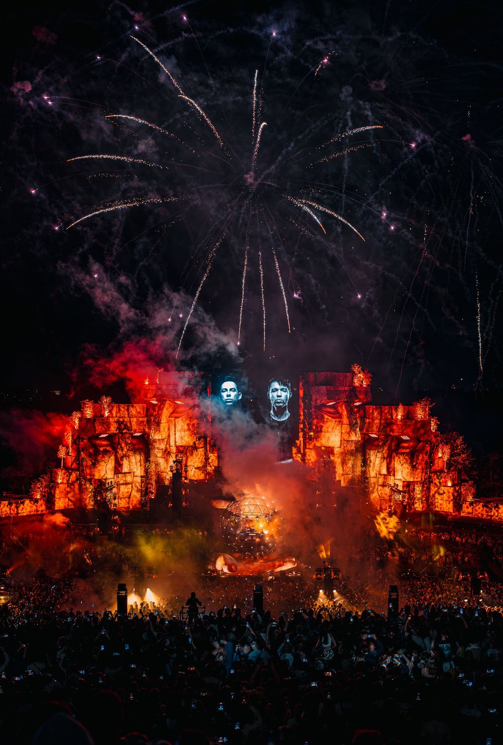 Spectacular fireworks explode over an ancient-themed festival stage with fiery pyrotechnics at night.