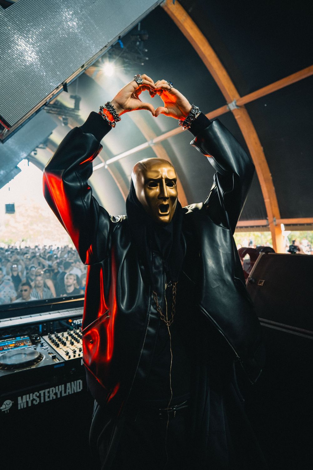 Masked DJ wearing gold face mask making heart shape with hands at Mysteryland music festival.