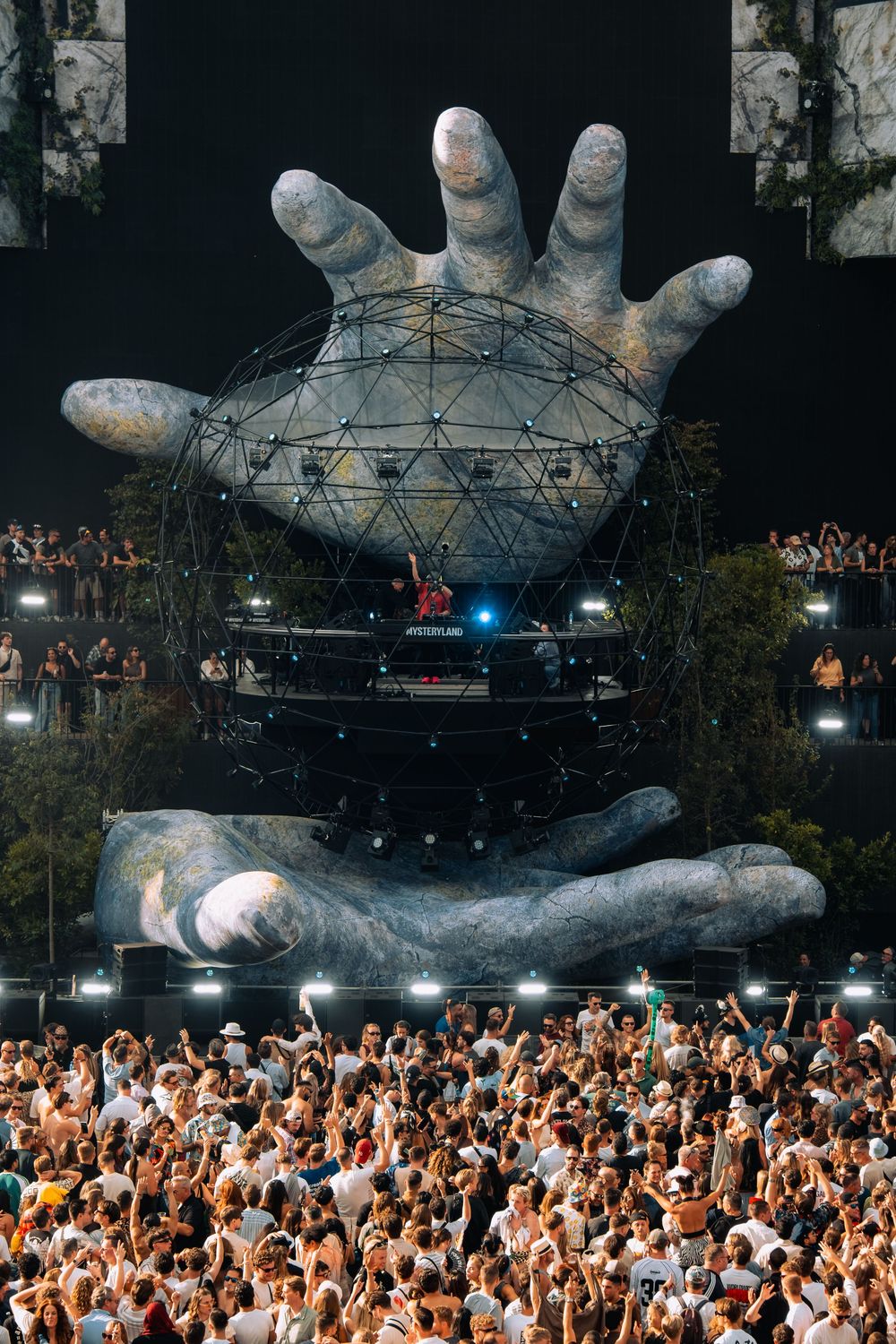 Massive crowd at Mysteryland festival facing a giant stone hands sculpture with geodesic dome stage.
