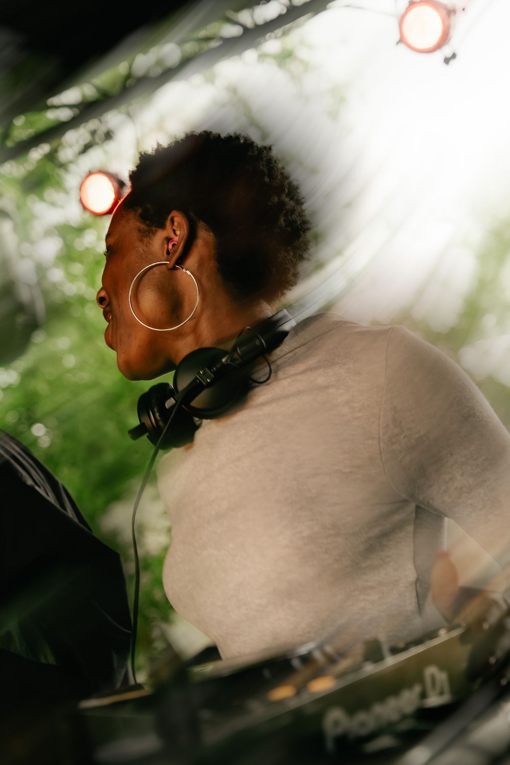 Female DJ with hoop earrings and headphones around neck performing at an outdoor music event with stage lights.