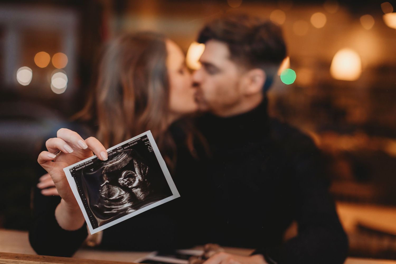 Couple kissing while holding ultrasound sonogram photo, pregnancy announcement in romantic bokeh setting.