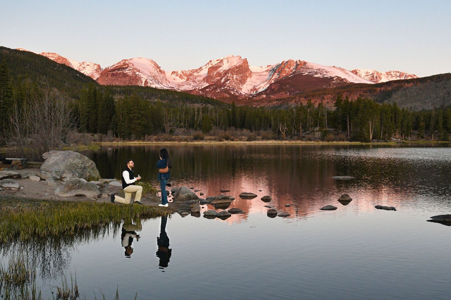 Man proposing to woman at Sprague Lake with Hallets peak glowing pink in the sunrise. Pixcapades | The Rocky Mountain Proposal Photographer