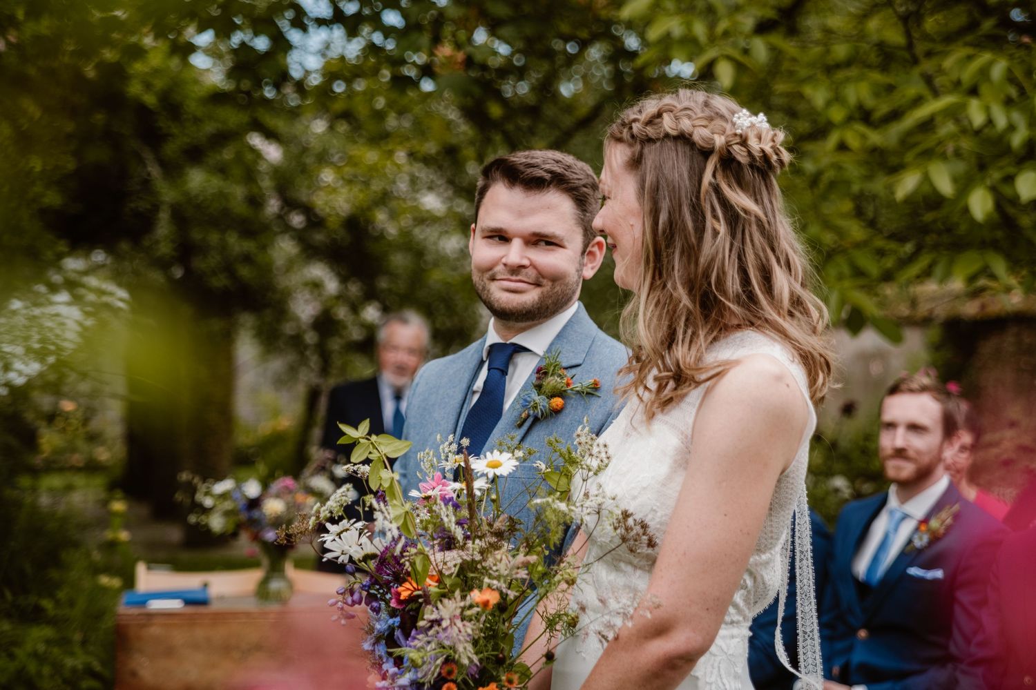 Groom in blue suit lifts laughing bride in white gown before vibrant pink rhododendron blooms in garden.
