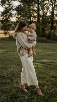A woman in neutral tones holds a baby outdoors at golden hour in a grassy field.