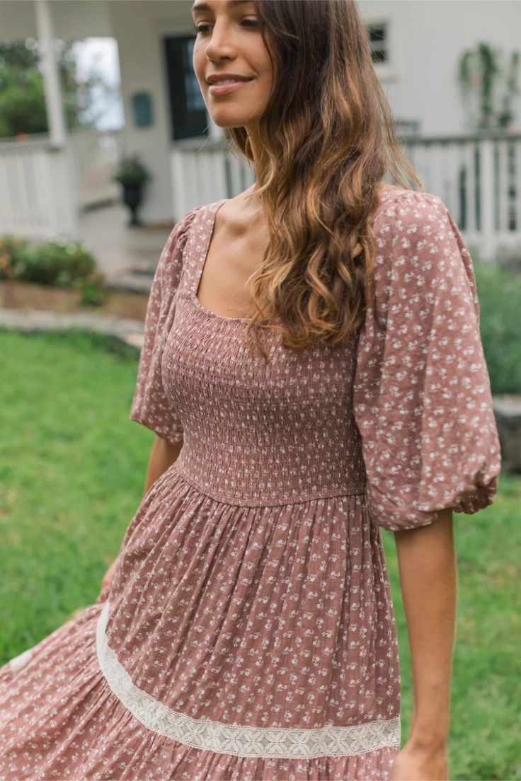 Woman wearing a mauve floral smocked bodice dress with puff sleeves and lace trim, posing outdoors.