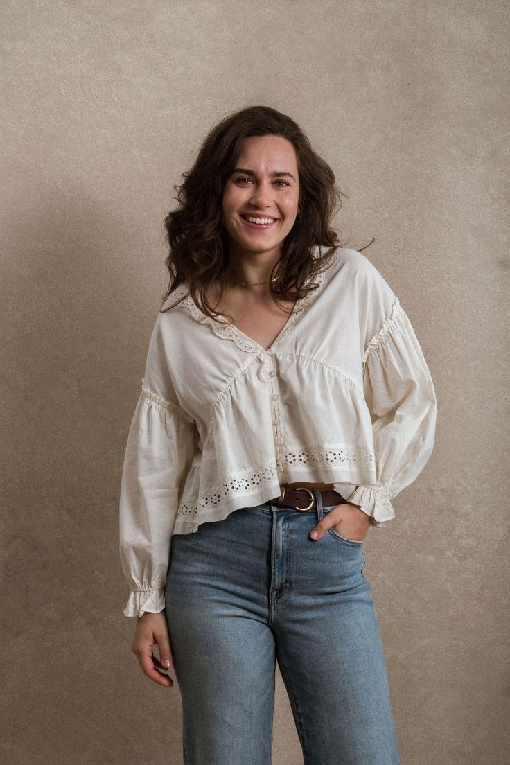 Woman smiling in white eyelet lace blouse with V-neck and blue jeans, posing against neutral background.