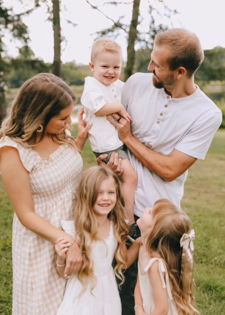 Young family of five posing outdoors, parents with three children in white outfits among green trees.