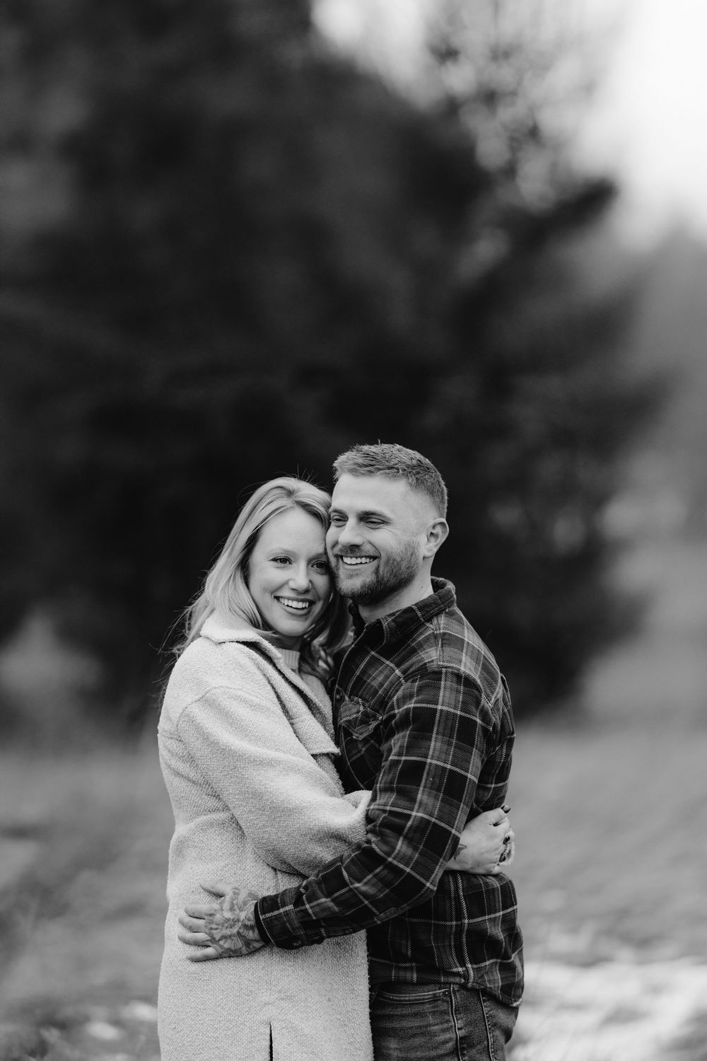 Black and white photo of a happy couple embracing and smiling outdoors among lush green trees.