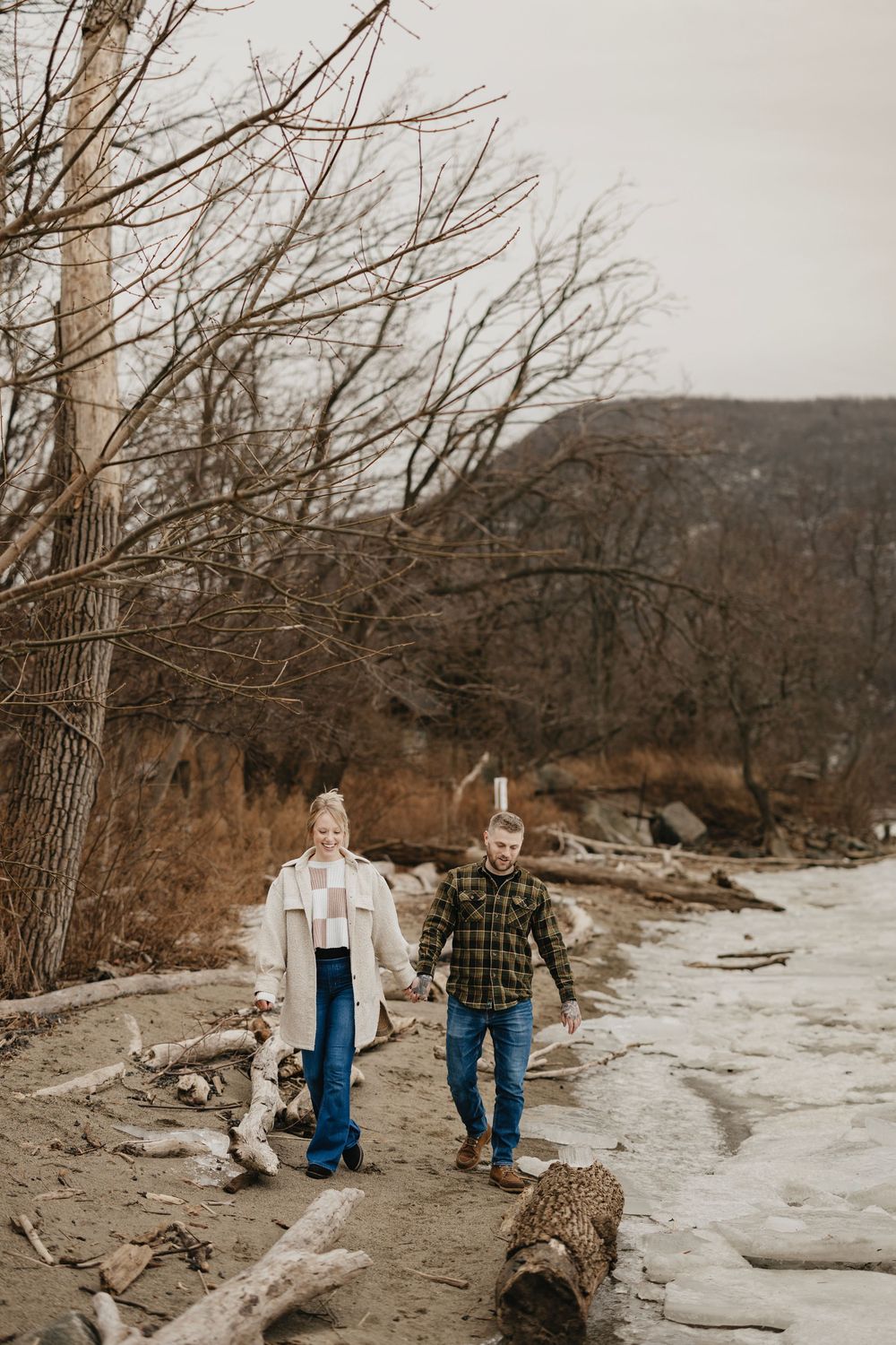 Couple walking hand in hand along a rocky winter shoreline surrounded by bare trees and driftwood.
