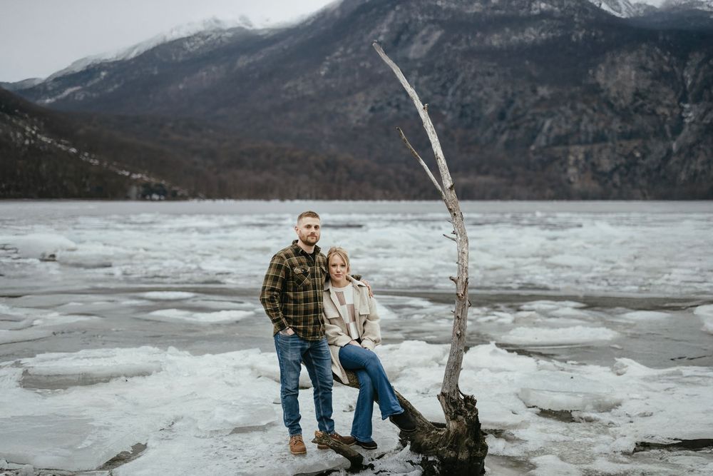 Couple posing on driftwood by a frozen mountain lake with icy shores and snow-capped peaks in the background.