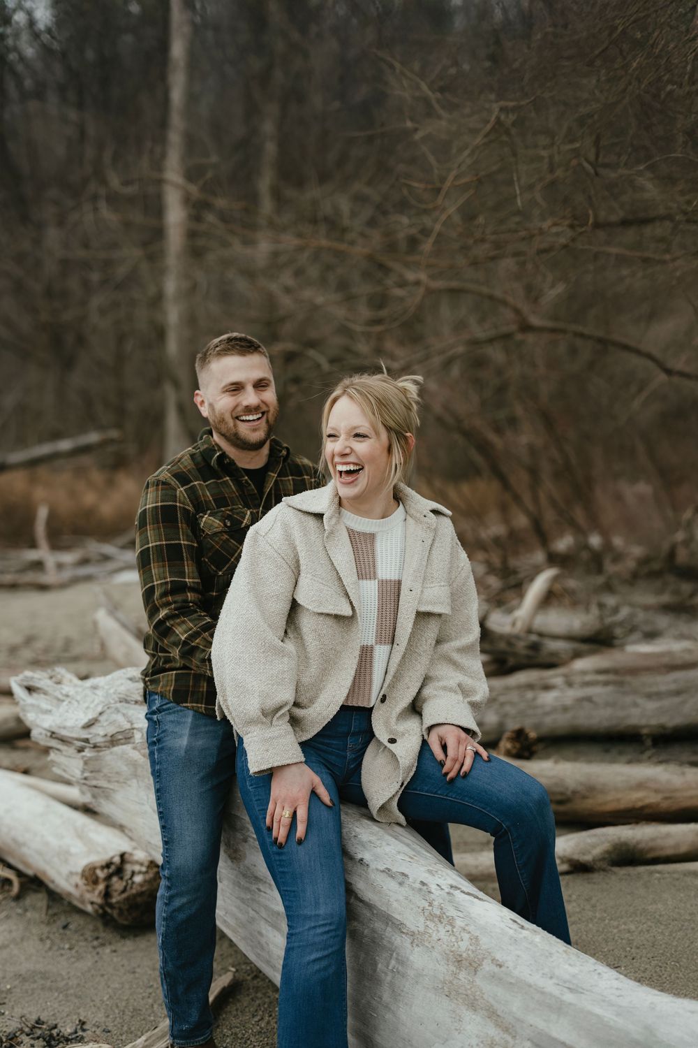 A laughing couple sits on driftwood logs in a forest, wearing casual fall outfits of flannel and cream jacket.