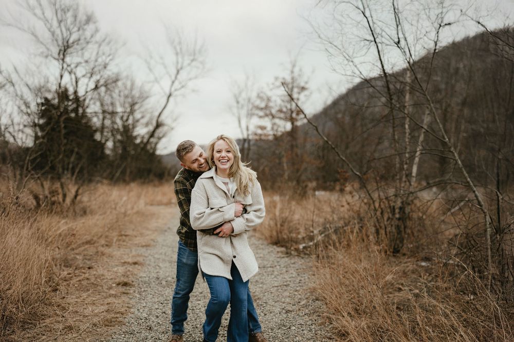 Couple laughing together on a rustic trail surrounded by golden grass and bare trees in autumn.