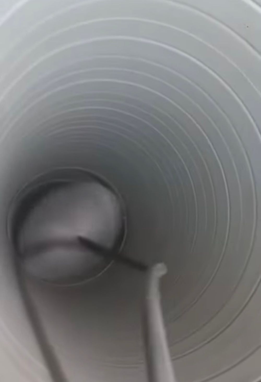 Close-up interior view of a gray corrugated plastic pipe with a round plug and metal rod visible inside.