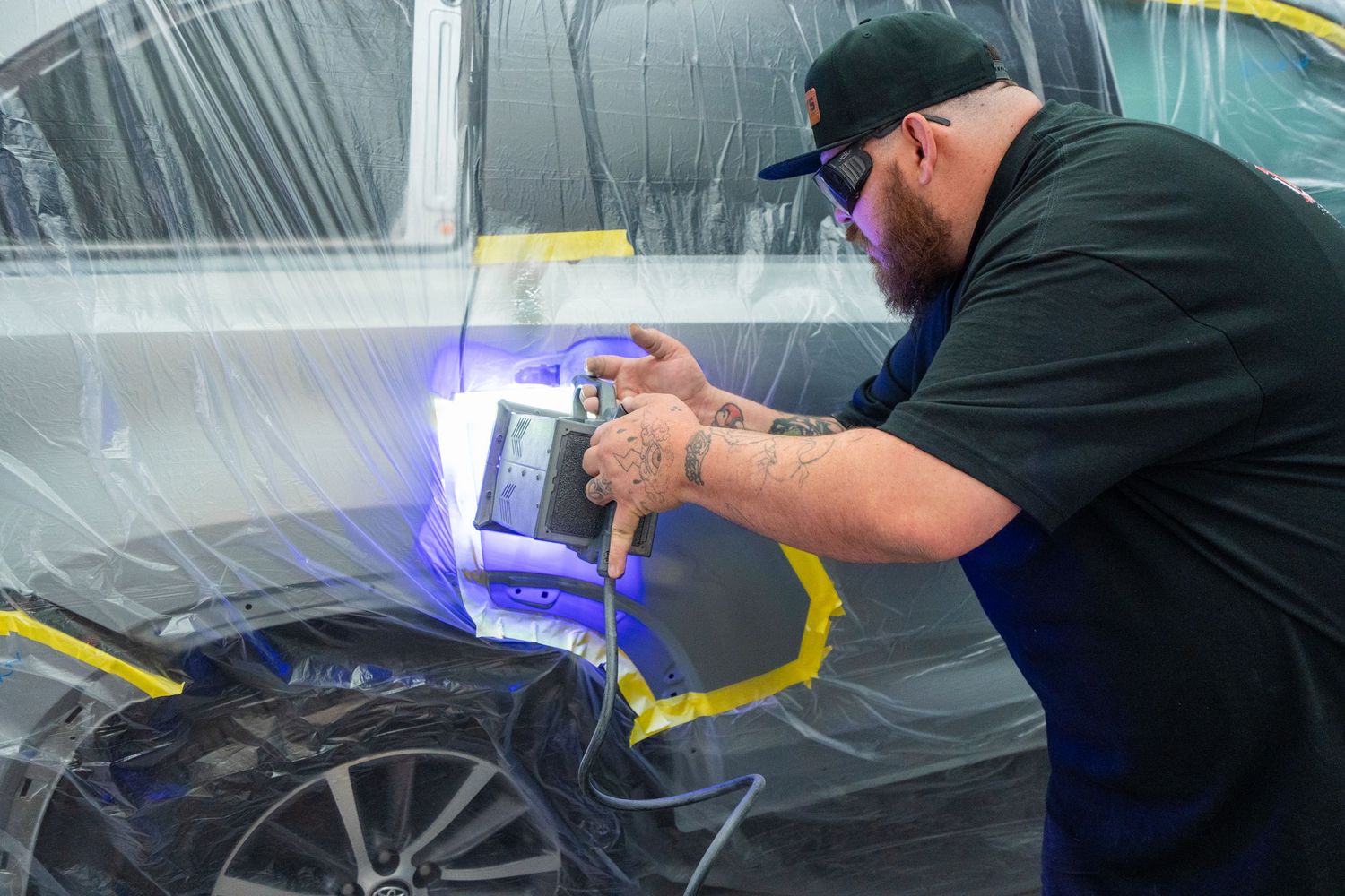 Technician using a paint thickness gauge on a dark car in a professional auto body shop.