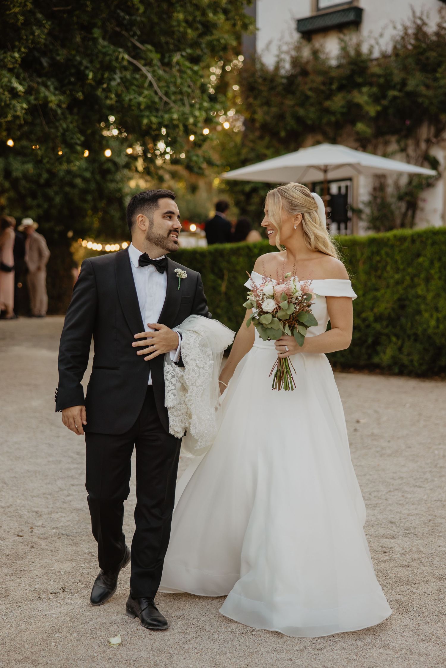 Bride in off-shoulder gown holding bouquet walks with groom in black tuxedo at elegant outdoor wedding venue.