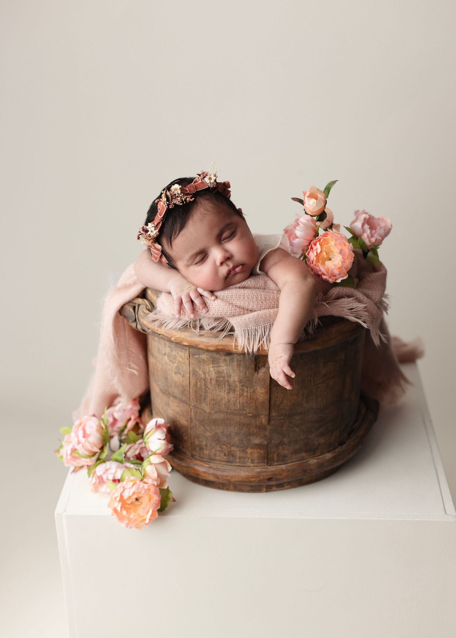 Sleeping newborn baby girl in wooden bucket with pink floral headband and peony flowers in soft studio setting.