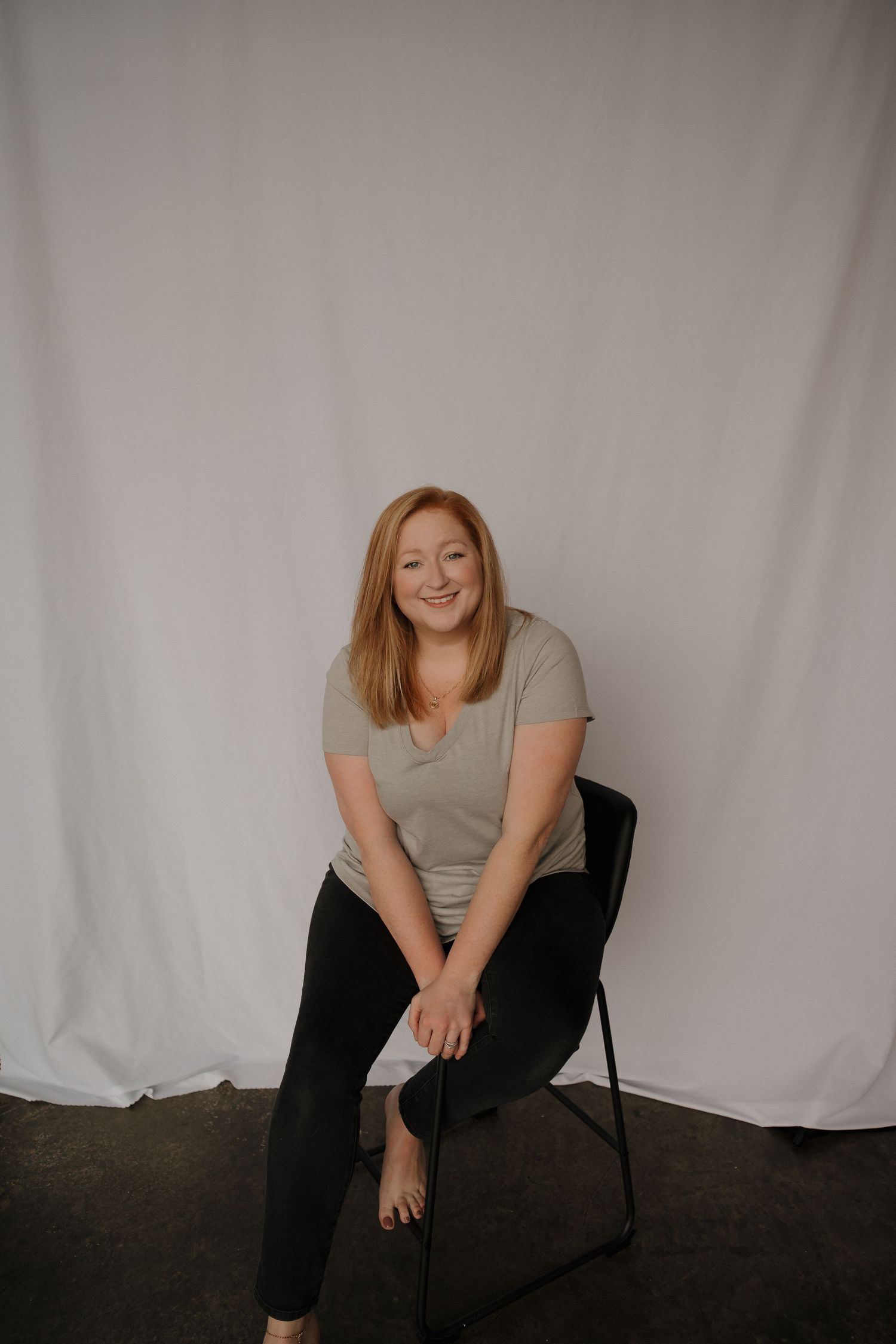 A smiling redhead woman sits relaxed on a black chair against a white fabric backdrop, wearing a sage top and black pants.