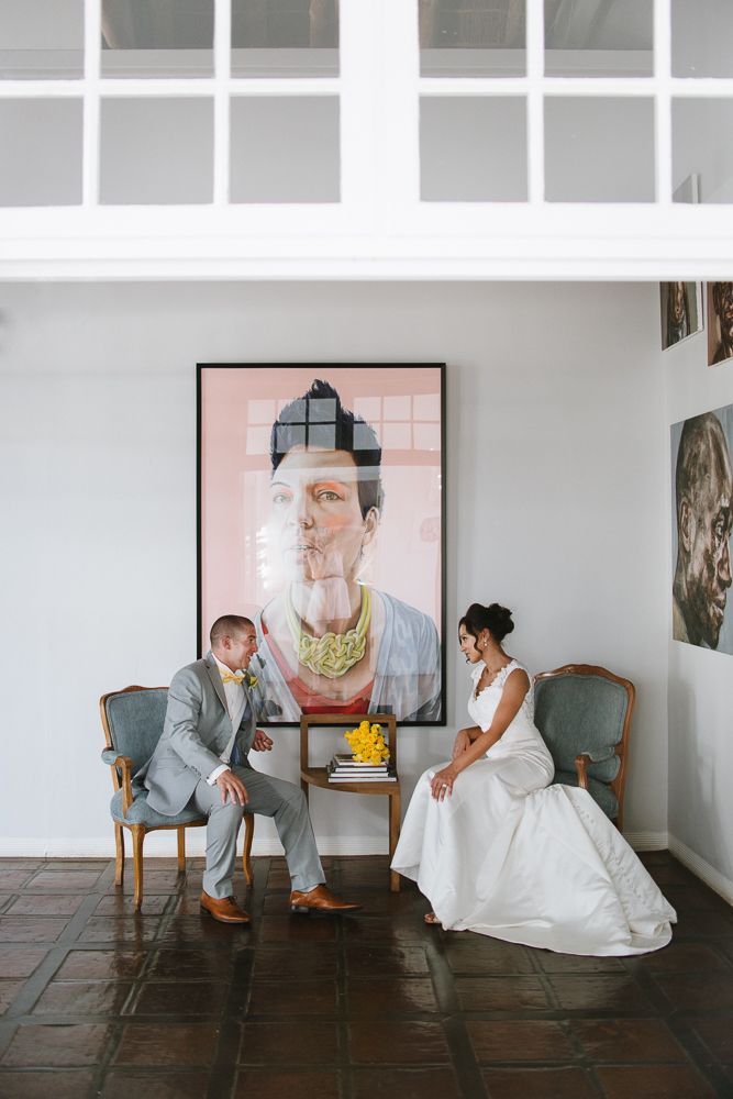 Bride and groom sit in vintage chairs before a large portrait painting in an elegant white gallery space.