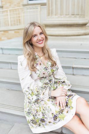 Smiling blonde woman in a white floral dress sitting on stone steps outdoors.