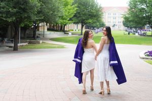 Two women in white dresses walk together on a brick path, one wearing a purple graduation gown, on a college campus.