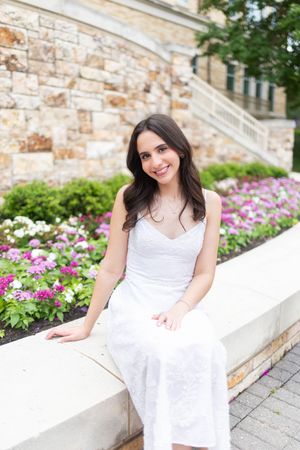 Young woman in white dress sitting on stone wall surrounded by colorful flowers and greenery, smiling outdoors.